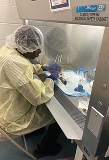 The photo on this page shows Dr. Alex Kayongo, a Ugandan man wearing a white hairnet and yellow coverall, working with samples in his lab.