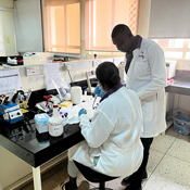 The photo on this page shows Dr. Alex Kayongo, a Ugandan man wearing a lab coat, consulting with a woman, similarly dressed in a lab coat. She sits and he stands at a desk in his laboratory