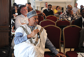 Shehu Usman Adamu holds a microphone as he asks a question from the seated audience at the 2023 Fogarty Bioethics Network Meeting. Other attendees listen in.