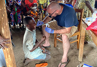 The photo shows Dr. Matthew Bramble, dressed in shorts and a t-shirt, using a swab to take a sample from the mouth of a child affected by konzo in Kahemba.