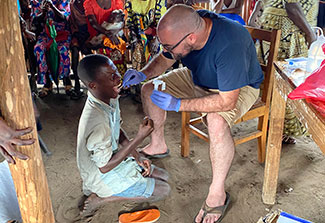 The photo shows Dr. Matthew Bramble, dressed in shorts and a t-shirt, using a swab to take a sample from the mouth of a child affected by konzo in Kahemba.