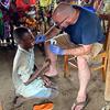 The photo shows Dr. Matthew Bramble, dressed in shorts and a t-shirt, using a swab to take a sample from the mouth of a child af