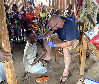 The photo shows Dr. Matthew Bramble, dressed in shorts and a t-shirt, using a swab to take a sample from the mouth of a child affected by konzo in Kahemba.