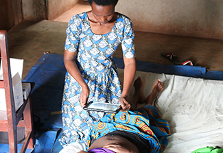 This photo shows a community health worker dressed in a blue and white dress using an tablet with an artificial intelligence–powered app to see whether a woman's surgical wound has become infected.