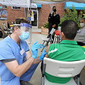 Dr. Michael Herce, wearing scrubs and protective mask, giving a shot in the arm to a man sitting in a chair 