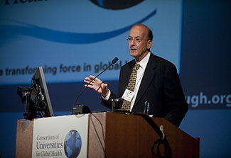 This photograph shows Dr. Roger Glass standing at a podium adorned with the Consortium of Universities for Global Health banner. He is addressing the annual meeting of CUGH on the NIH Campus at the Natcher Building.