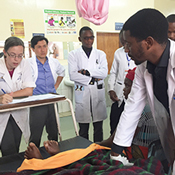 In the photograph a group of trainee doctors, all wearing white lab coats, circle a patient’s bed. Dr. Deanna Saylor writes in the patient’s chart as Dr. Mashina describes the patient’s condition.