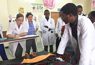 In the photograph a group of trainee doctors, all wearing white lab coats, circle a patient’s bed. Dr. Deanna Saylor writes in the patient’s chart as Dr. Mashina describes the patient’s condition. 