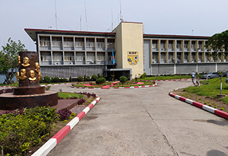 This photo shows the exterior of and drive up to the School of Public Health building at Kinshasa University. A statue featuring likenesses of various professors is in the foreground to the left.