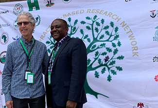 In the photo on this page, Drs. Gregory Aarons and Eche Ezeanolue pose for a photo together in front of the white Nigeria Implementation Science Alliance banner. Aarons wears sunglasses, black pants and a blue striped shirt. Ezeanolue wears an open collar shirt beneath a dark suit.  