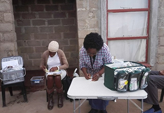 A health worker sits at a small folding table writing on a form, with an open medical kit bag containing supplies beside her. Another woman sits nearby holding paperwork, and a wire basket of medical supplies rests on a stand to the left. The setting appears to be an outdoor clinic or community health outreach site next to a brick building