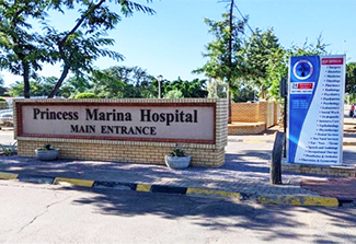 The photo shows the main entrance to the Princess Marina hospital, a district hospital in Botswana, on a pleasant sunny day. Surrounding the hospital are green trees and shrubs.