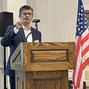 Junaid Razzak standing at a podium, speaking through a microphone with an American flag in the background