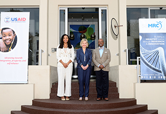 From left: Paloma Adams-Allen (Deputy Administrator of the USAID) , Glenda Gray (SAMRC President and CEO) and Reuben Brigety (United States Ambassador to South Africa) stand outside SAMRC flanked by vertical banners for USAID to the left and SAMRC to the right. 