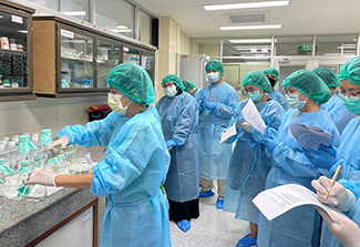 A group of healthcare workers wearing teal surgical gowns, caps, and masks gather in a hospital or lab room, with one person handling supplies at a counter while others hold clipboards and papers, some taking notes, in front of glass-fronted cabinets stocked with medical supplies.