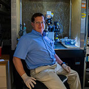 Yuriy Usachev, wearing glasses, a light blue shirt and tan pants, sits next to a microscope and computer screens at his lab at the University of Iowa.
