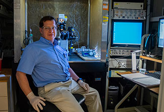 Yuriy Usachev, wearing glasses, a light blue shirt and tan pants, sits next to a microscope and computer screens at his lab at the University of Iowa.