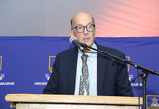 Roger Glass, Wearing a dark blue suite and light blue shirt, stands at a podium speaking into a microphone at the 2022 AFREHealth Symposium.