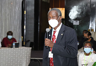 Francis Omaswa, holding a microphone and wearing a dark suit, red tie, and mask, speaks at the 2022 AFREHealth Symposium.