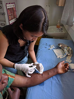 Dr. Aileen Chang wearing gloves uses needle to administer to wounds on patient’s arm resting on a covered table in a clinic
