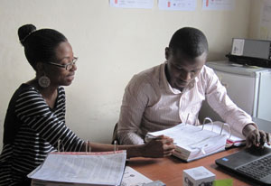 Dr. Juliana Anyanwu seated at a desk reviews large binders full of data, man seated next to her enters data on a laptop