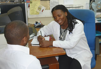Dr Akwi Asombang in white coat with stethoscope seated at desk takes note on pad while speaking with a seated patient