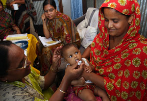 Woman wearing head scarf holds baby who receives oral vaccine administered by another woman in crowded clinic