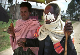 Man with white bandages taped over both eyes holding walking stick walks on dirt road, holding arm of a woman holding umbrella