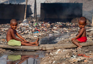 Two young, shirtless boys seated outdoors surrounded by garbage and rubbish, and stagnant water, background air is hazy