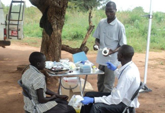 Two male researchers interview and collect samples from young study participant outdoors in shade next to folding table