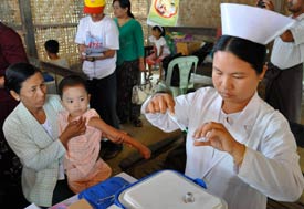 Burmese nurse in foreground prepares syringe, woman seated holds out arm of young child