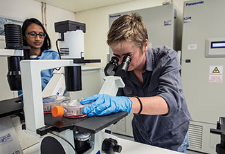 Dr. Penny Moore in lab looks at sample using microscope while Dr. Jinal Bhiman observes