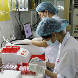 Two researchers work in lab wearing white coats, gloves, masks on face and hair coverings.