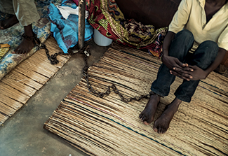 Overhead shot of a man sitting from the neck down, hands resting on over his knees, his ankle cuffed and chained to another man's ankle cuff.