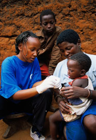 Heath care worker wearing latex gloves works with young child, who sits in its mother's lap, another child looks on