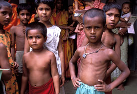 Two small, thin, shirtless boys in foreground, in background many other children stand, women hold toddlers and infants