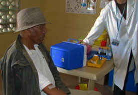 Older man seated in clinic holds out arm, bandage in crook of elbow, clinician in background arranges vials on table
