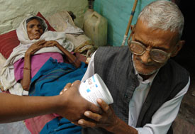 Older, thin, sickly woman lies on cot in background, older man at the foot of the cot receives container from someone standing o