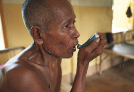 Older man uses inhaler, shirtless, bare floored clinical setting with empty bedframes in background