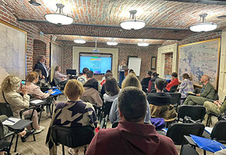 Dr. Tetiana Nickelsen stands at a podium with a screen showing a slide with the title “Cognitive Processing Therapy for PTSD.” Students are seated on folding chairs in an underground bunker in Lviv, Ukraine.