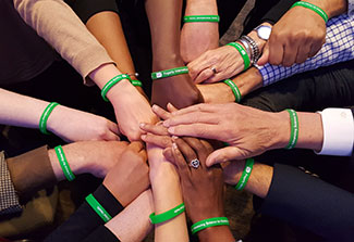 Hands join in the middle of a huddle pictured from above, all wearing bright green Fogarty wristbands.