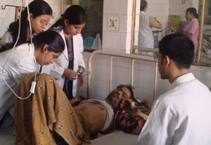 A weak looking man lies in metal frame hospital bed, medical workers examine him, 3 female standing, 1 male seated
