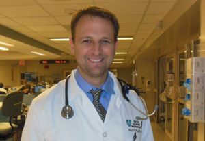 Dr Paul drain smiles at camera in hall of a hospital, wearing white doctor's coat, stethoscope around neck