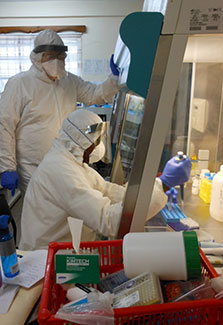 Lab worker in protective clothing and mask works with samples under a hood, another worker observes