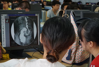 Two women with backs to camera review a medical image on a monitor in a crowded computer lab