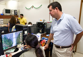 Woman seated works on computer, man stands behind her looking on, in busy lab
