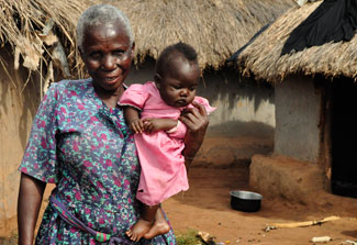 Older woman holds baby outdoors, thatched huts in the background