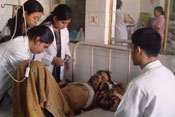 A weak looking man lies in metal frame hospital bed, medical workers examine him, 3 female standing, 1 male seated