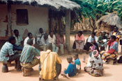 Large group of adults and children, seated on stools and dry dusty ground in circle, huts in background