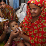 Photo courtesy of PATH: Woman wearing head scarf holds baby who receives oral vaccine administered by person off camera